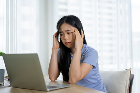 A stressed and confused young Asian woman in casual clothes sits at a table in her room looking at her laptop screen with a doubtful face, worried about her project, working with pressure.の写真素材