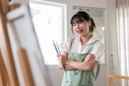 A cheerful and creative young Asian female artist in an apron smiles at the camera, stands with her arms crossed, and holds paint brushes in her hand in her home studio workshop.の写真素材