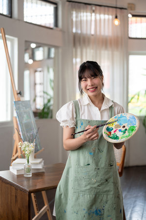 A cheerful and beautiful young Asian female artist in an apron holding a paint brush and a color...