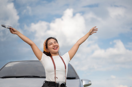 A cheerful and beautiful young Asian woman raises her hands and stands in front of her car, feeling carefree and relaxed after arriving at her destination by car on her summer vacation.の写真素材
