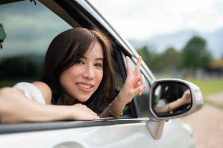 A beautiful young Asian woman is showing a peace hand sign, smiling at the camera from the car window, and enjoying her road trip in the summer. road trip, summer vacation, travelの写真素材