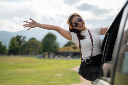 A happy and cheerful young Asian woman in sunglasses is enjoying the wind outside the car, having...