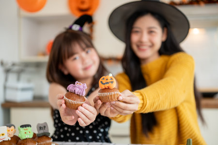Selective focus image, A happy and cute young Asian girl and her mom in Halloween costumes are enjoying decorating Halloween cupcakes in the kitchen together. Spooky season, family timeの写真素材