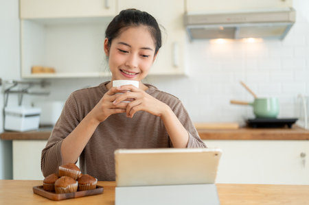 A happy and beautiful young Asian woman is working from home, sipping her morning coffee and using her digital tablet at a table in the kitchen. People and technology conceptsの写真素材