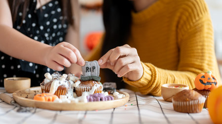 Close-up hand image of a young Asian girl and her mom are decorating Halloween cupcakes in the kitchen together. Spooky season, family timeの写真素材