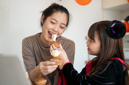 A caring and beautiful young Asian mom enjoys eating Halloween cupcakes and celebrating Halloween with her little daughter at home.の写真素材