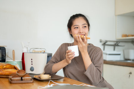 A beautiful and happy young Asian woman is enjoying her toast and coffee in the kitchen, having an easy breakfast at a dining table in the kitchen.の写真素材