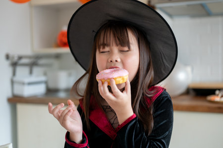 A cute and happy young Asian girl in a Halloween costume enjoys eating a Halloween cupcake in the kitchen, celebrating spooky season at home.の写真素材