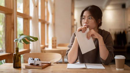 A pensive, thoughtful young Asian woman sits at a table in a beautiful, cosy coffee shop, looking...