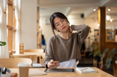 A tired and overworked young Asian woman is massaging her neck while working remotely at a coffee shop. Student, freelancer, businesswomanの写真素材