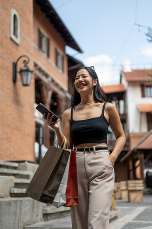 A portrait of an attractive and cheerful Asian woman in trendy clothes enjoys shopping in the city, walking while holding her phone and shopping bags. Urban lifestyle, city girl, shopping spreeの写真素材