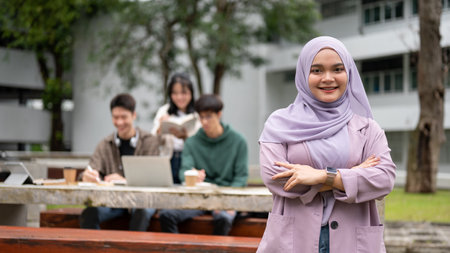 A smiling and charming young Asian-Muslim female university student in a purple hijab stand with her...
