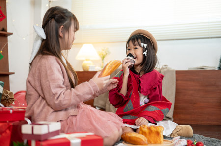 Two joyful and pretty young Asian girls in cute Christmas dresses are enjoying eating Christmas sweets in the living room and celebrating Christmas at home together. Happy family, siblings, Christmasの写真素材