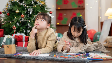 Two cute and happy young Asian girls are lying on the living room's floor near the Christmas tree and enjoying drawing and coloring together.の写真素材