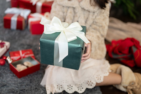 A cute young girl in a cute dress is holding a Christmas present gift box while sitting under the Christmas tree in the living room, celebrating Christmas at home. Close-up imageの写真素材