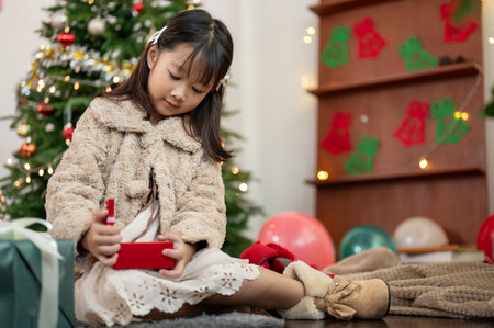 A cute and happy young Asian girl in a cute dress is opening a Christmas gift in the living room, surprised with her Christmas present and celebrating Christmas at home.の写真素材