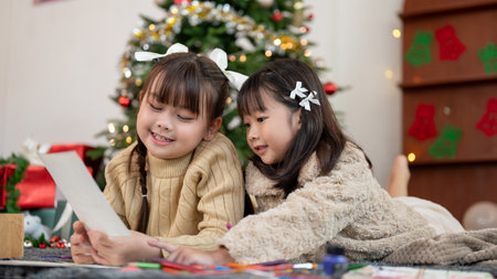 Two adorable and cheerful young Asian girls are having fun, enjoying talking and making Christmas cards while lying on the living room's floor together. Merry Christmas, kid leisure, art, inspirationの写真素材