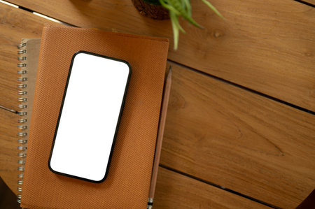 A white-screen smartphone mockup on a book on a wooden table. study table, office desk, minimal workspace top view imageの写真素材