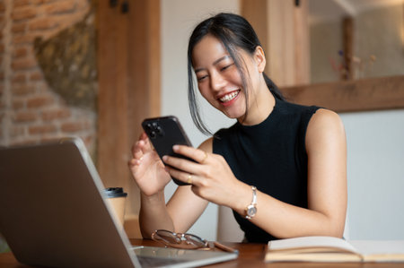 A cheerful and beautiful young Asian woman is laughing and enjoys chatting with her friends or watching funny videos online on her smartphone while sitting in a coffee shop.の写真素材