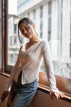A portrait of an attractive Asian woman in casual clothes is looking at the camera and standing against the window indoors.の写真素材