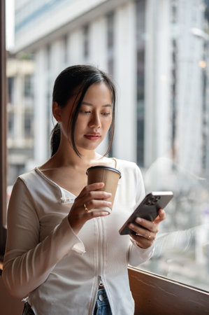 A portrait of a beautiful Asian woman using her smartphone, reading online blogs or messages, and holding a coffee cup while standing by the window.の写真素材