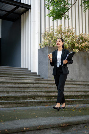 A beautiful and confident millennial Asian businesswoman in a formal business suit is holding a coffee cup and document binders while walking down the stairs in front of the corporate building.の写真素材