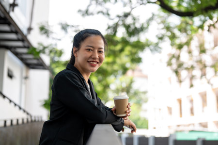 A cheerful and beautiful millennial Asian businesswoman in a black suit is relaxing outdoors during her coffee break. Business people conceptの写真素材
