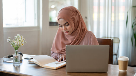 A beautiful and concentrated young Asian-Muslim woman is reading a book while working remotely at a cafe on the weekend.の写真素材