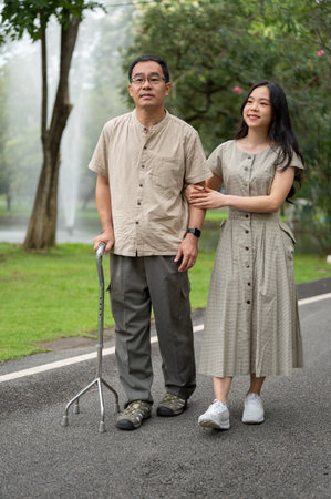 A smiling and lovely young Asian daughter and her happy senior dad with a walking stick are walking in the park in the morning together.の写真素材