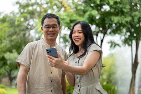 A cheerful and lovely young Asian daughter is laughing, showing a funny clip on her smartphone to her dad while relaxing in the green park in the morning together. Happy family conceptの写真素材