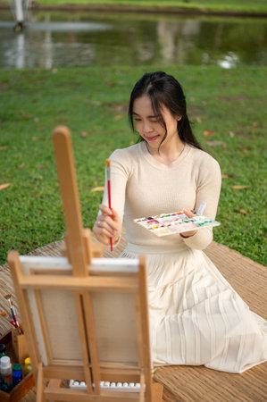 A portrait of an attractive and creative young Asian woman is painting or drawing on a canvas while relaxing in a green park.の写真素材