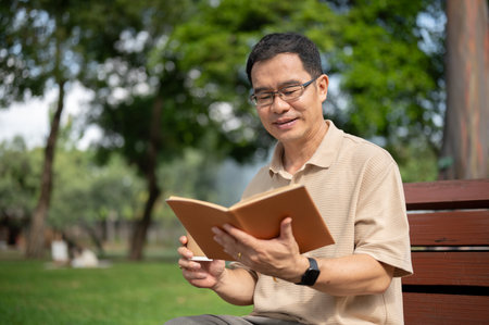 A happy senior retired Asian man wearing eyeglasses is reading a book and having morning coffee while relaxing on a bench in a green park. Happy retirement lifestyle conceptの写真素材