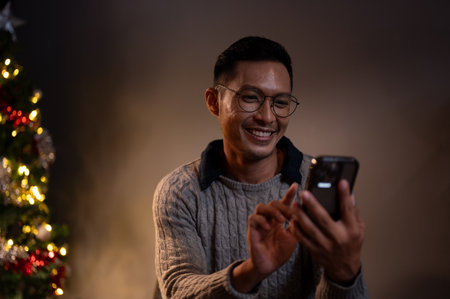 A cheerful Asian man in a cosy sweater is chatting with his friends or sending Christmas messages to his family while relaxing in his living room on a Christmas night.の写真素材