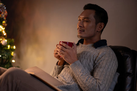 Side view image of a relaxed and calm Asian man in a cosy sweater, eyes closed, smelling his coffee, enjoying his coffee or hot cocoa in a living room on his Christmas night at home alone.の写真素材