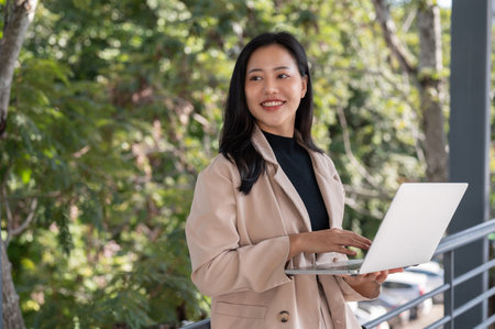 An attractive millennial Asian businesswoman working on her laptop while standing on a skywalk in the city, responding to urgent email.の写真素材