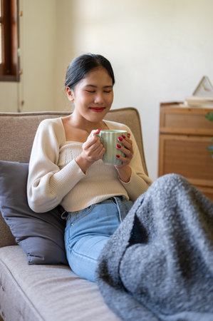 A happy, beautiful Asian woman in casual clothes is smelling her coffee, enjoying her morning coffee on a couch in her minimalist living room. Domestic life conceptの写真素材