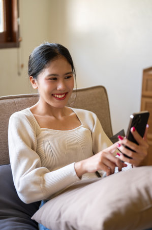 A happy and beautiful Asian woman enjoys watching videos online or chatting with her friends on her smartphone while relaxing on a couch in her living room.の写真素材