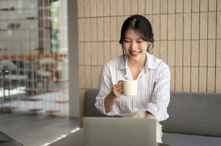 A beautiful young Asian woman is sipping coffee and working on her laptop at a table in a modern coffee shop. Urban life, lifestyleの写真素材