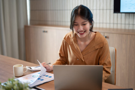 A smart and beautiful young Asian businesswoman or female entrepreneur is examining business financial reports, using her laptop and working at her desk in a modern office.の写真素材