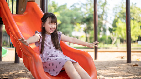 A playful and cheerful young Asian girl is having fun on summer, playing a colorful slide in an outdoor playground in the park.の写真素材