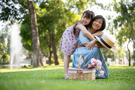 A happy senior Asian grandmother and her cute little girl granddaughter are hugging, enjoying tender moments, and piggy-back cuddling while having fun in the green park together.の写真素材