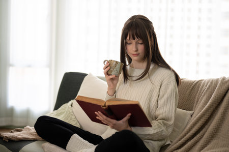 A pretty and relaxed young caucasian woman in a knitted sweater is reading a book and sipping coffee...