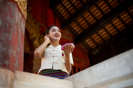 A charming and smiling Thai-Asian woman in a traditional dress with a garland in her hand is visiting a beautiful temple on a Buddhist holy day, looking away from the camera. Thai-culture conceptの写真素材