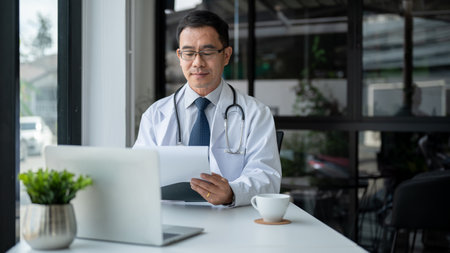An experienced Asian senior male doctor in a white gown is examining medical cases on report at his desk in the office, working in a hospital. Health care and career conceptsの写真素材