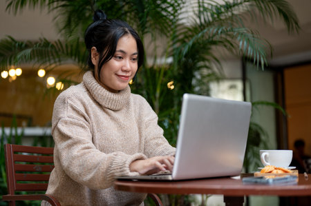 A beautiful young Asian woman in a cosy knitted sweater is working on her laptop computer at an outdoor table, working remotely at a cafe. Urban lifestyle conceptの写真素材