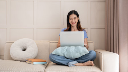 Young pretty Asian woman using laptop computer while sitting on the sofa in her living room. Life...