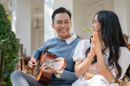 Cheerful and happy young Asian couple enjoying playing guitar and picnic at home backyard together. bonding, love, relationship conceptの写真素材