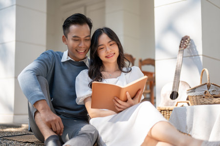 Lovely sweet young Asian couple enjoying picnic on a sunny day at home together, reading a book, having fun , enjoying talking.の写真素材