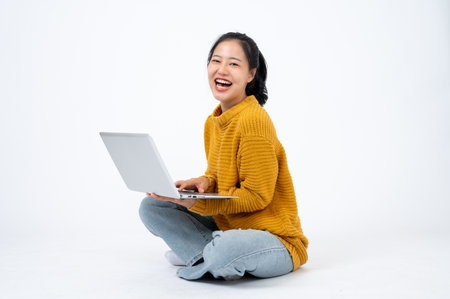Young happy and beautiful Asian woman in casual clothes sits on the floor on an isolated white background with her laptop computer. scrolling on websites, responding to emails, enjoys online shoppingの写真素材