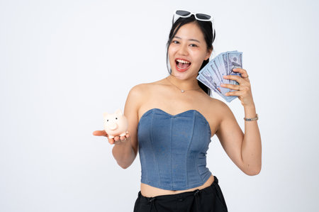 A beautiful and positive Asian woman in trendy clothes is holding a piggy bank and cash on an isolated white background. financial, banking, shopping, savingの写真素材
